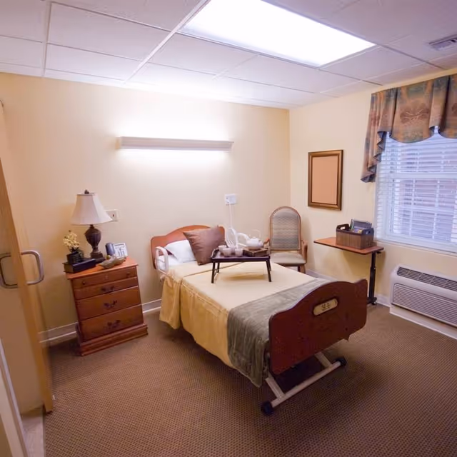 A small, neatly arranged bedroom in a senior living facility with a single bed covered in beige and gray bedding. A tray with a tea set is placed on the bed. Next to the bed is a wooden nightstand with a lamp, phone, and decorative items. A chair is positioned against the wall near a window with patterned curtains. The room has beige walls, a carpeted floor, and a wall-mounted air conditioning unit below the window.