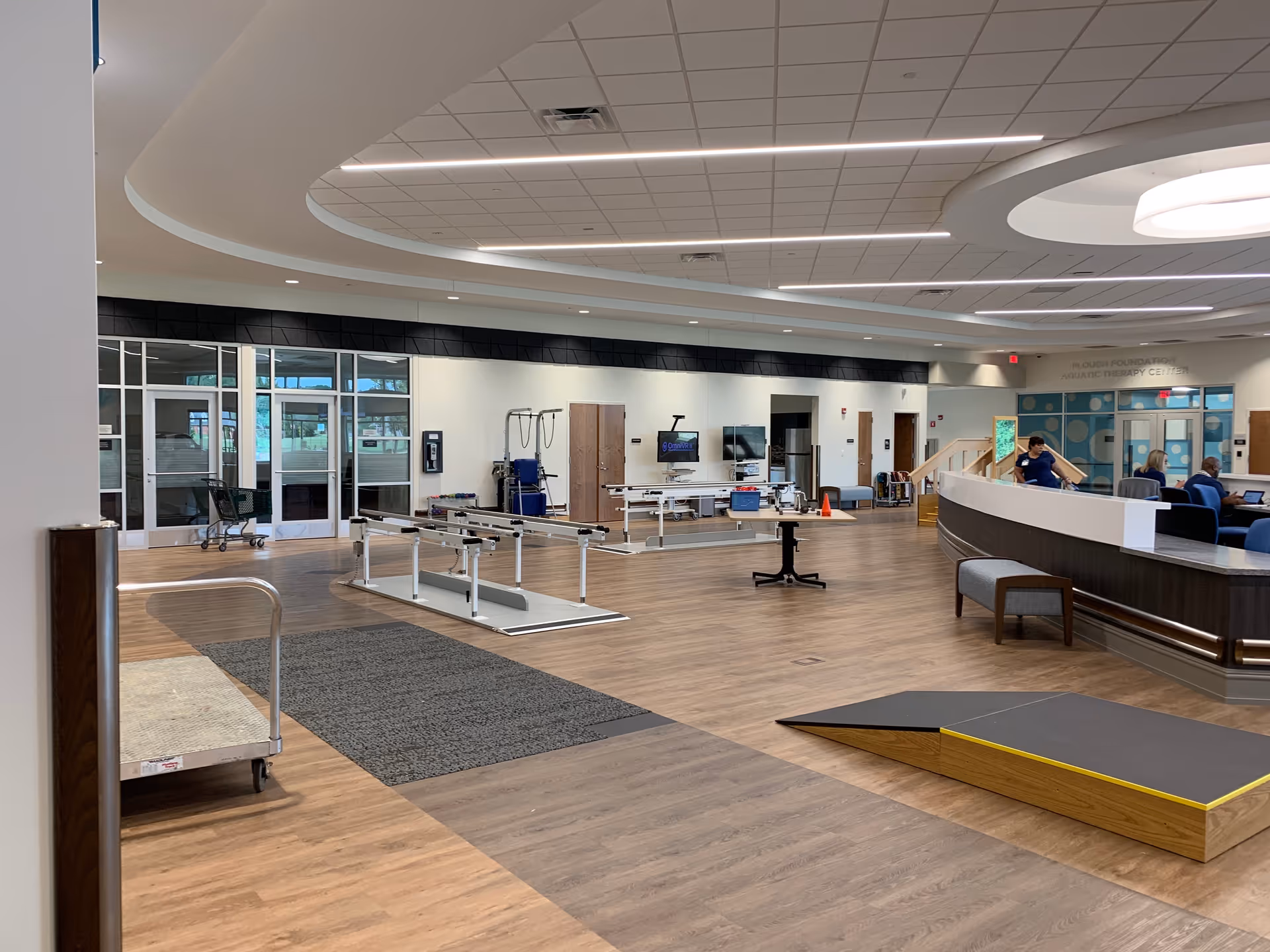 A spacious rehabilitation therapy room with wooden flooring and modern ceiling lights. The room features parallel bars for physical therapy, a ramp, and various therapy equipment. There is a curved reception desk with people seated behind it, and large glass doors and windows in the background.