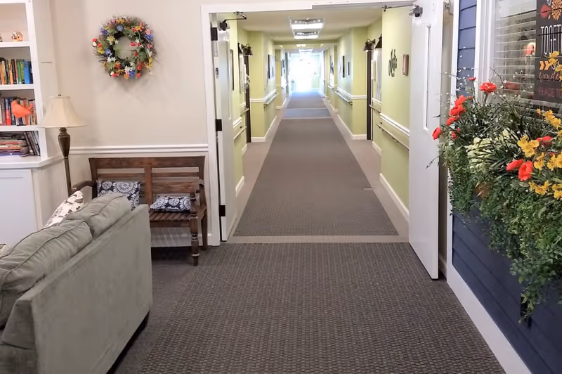 View of a hallway in a senior living facility with a gray carpet leading to a bright exit. On the left side, there is a green couch, a wooden bench with cushions, a floor lamp, and a bookshelf. A colorful wreath hangs on the wall above the bench. On the right side, there is a window with a flower box filled with red and yellow flowers and greenery.