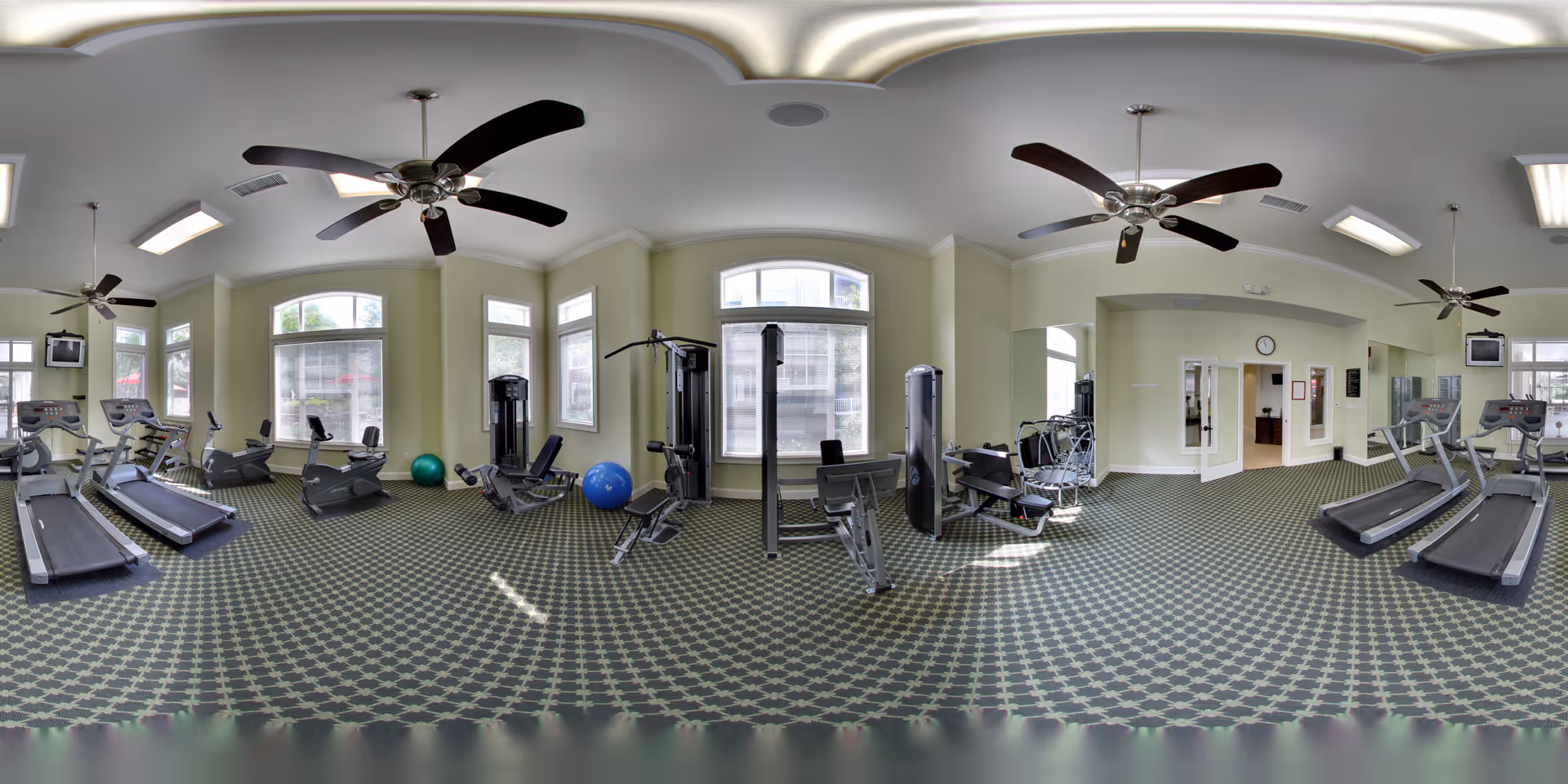 A spacious fitness room with multiple exercise machines including treadmills, stationary bikes, and weight machines. The room has large windows allowing natural light, ceiling fans, and patterned carpet flooring.