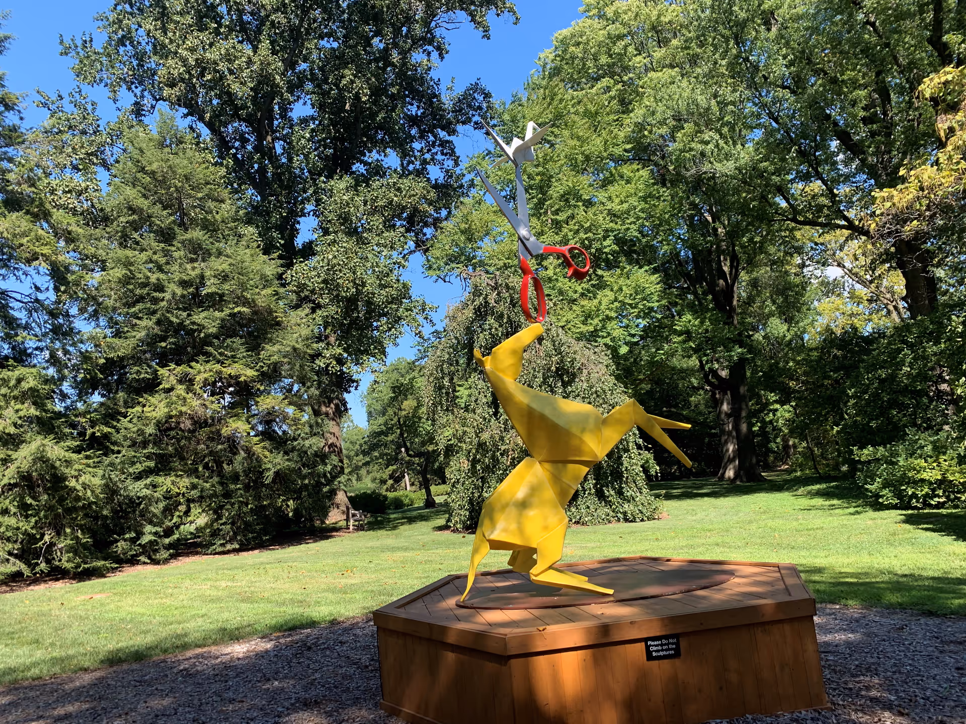 A yellow origami-style rabbit sculpture standing on its hind legs on a wooden platform in a park. The rabbit is holding a large pair of scissors with red handles in its mouth. The background features green trees and a clear blue sky.