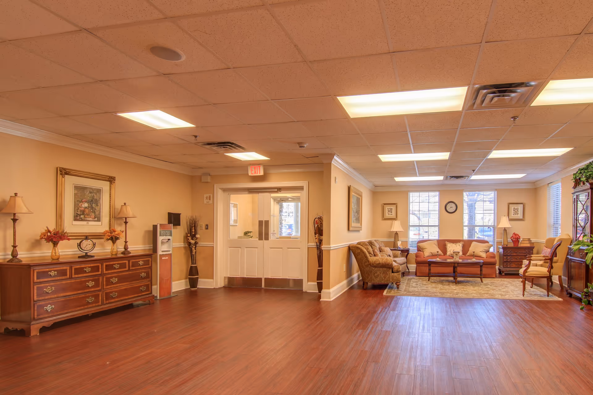 Spacious assisted living lobby with sofas, armchairs, wooden floors, and decorative furniture under a drop ceiling.