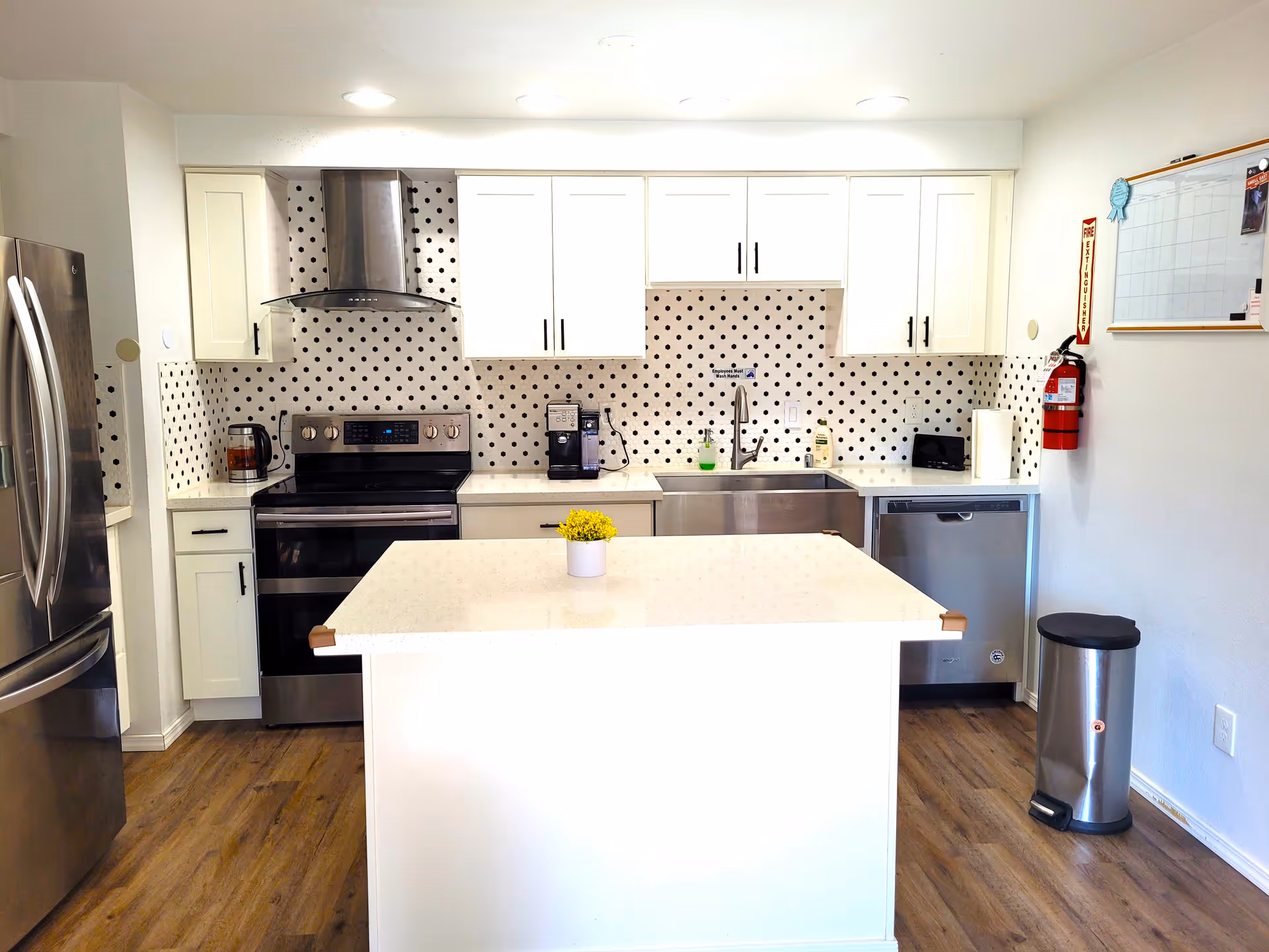 A modern kitchen with white cabinets, a stainless steel refrigerator, stove, dishwasher, and a central island with a small yellow flower pot. The backsplash features a white background with black polka dots. There is a fire extinguisher mounted on the wall next to a whiteboard calendar, and a stainless steel trash can on the right side.
