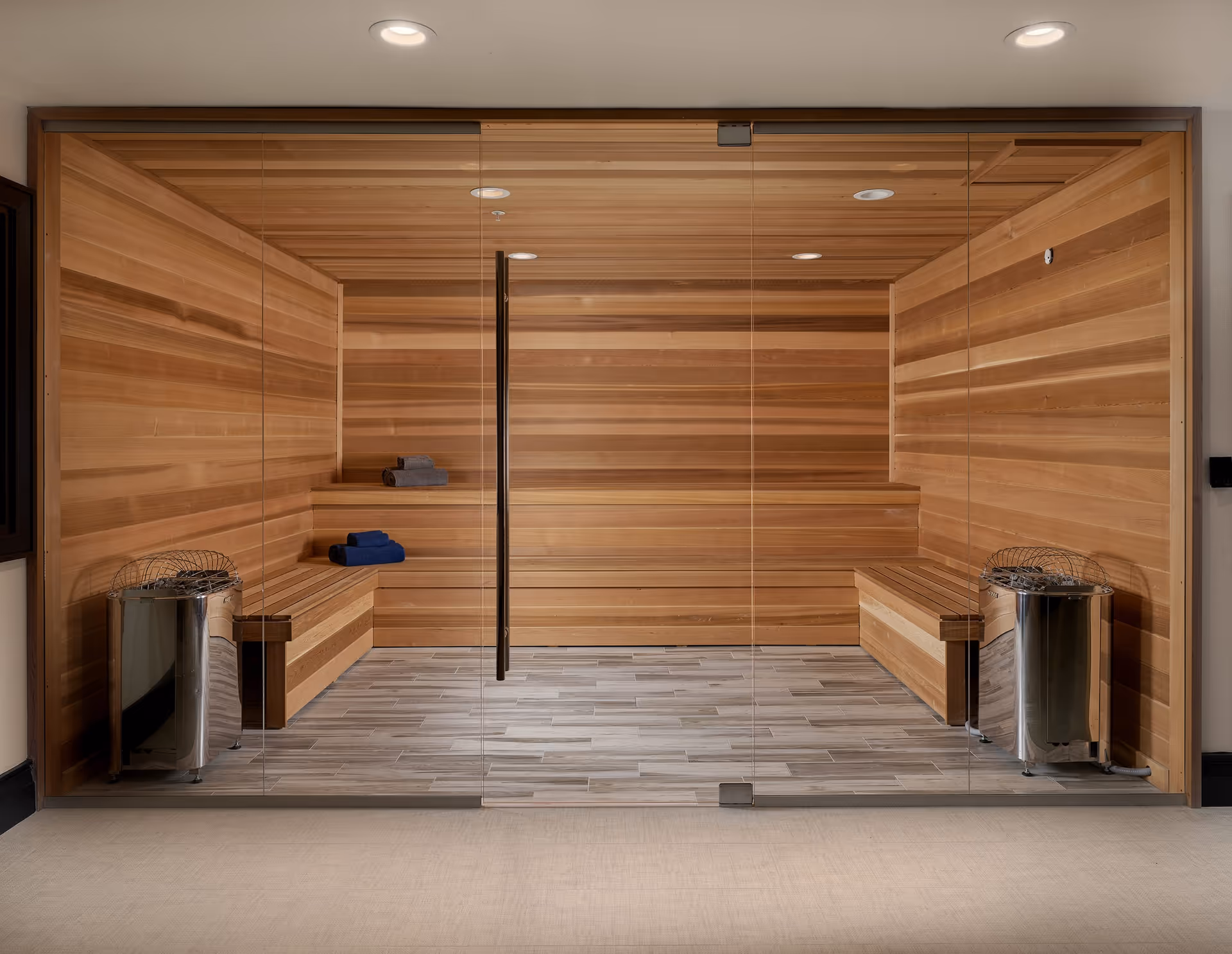 Interior view of a wooden sauna room with glass doors, wooden benches along the walls, two metal sauna heaters on each side, and folded towels placed on the benches.