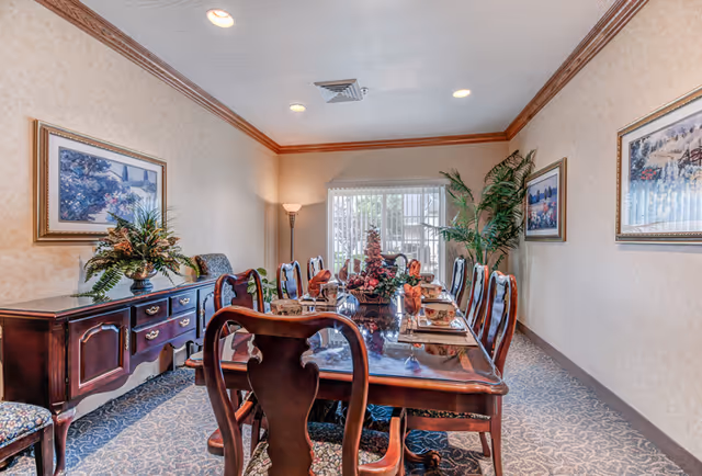 Formal dining room with a long polished wooden table set for a meal, matching chairs, sideboard, artwork, and potted plants.