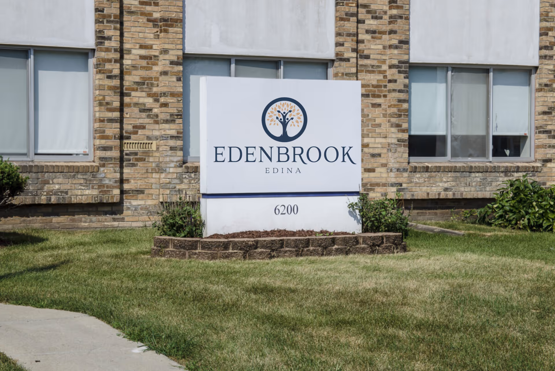 A white sign with the logo and text 'EDENBROOK EDINA' and the number 6200 in front of a brick building with windows and green grass surrounding the sign.