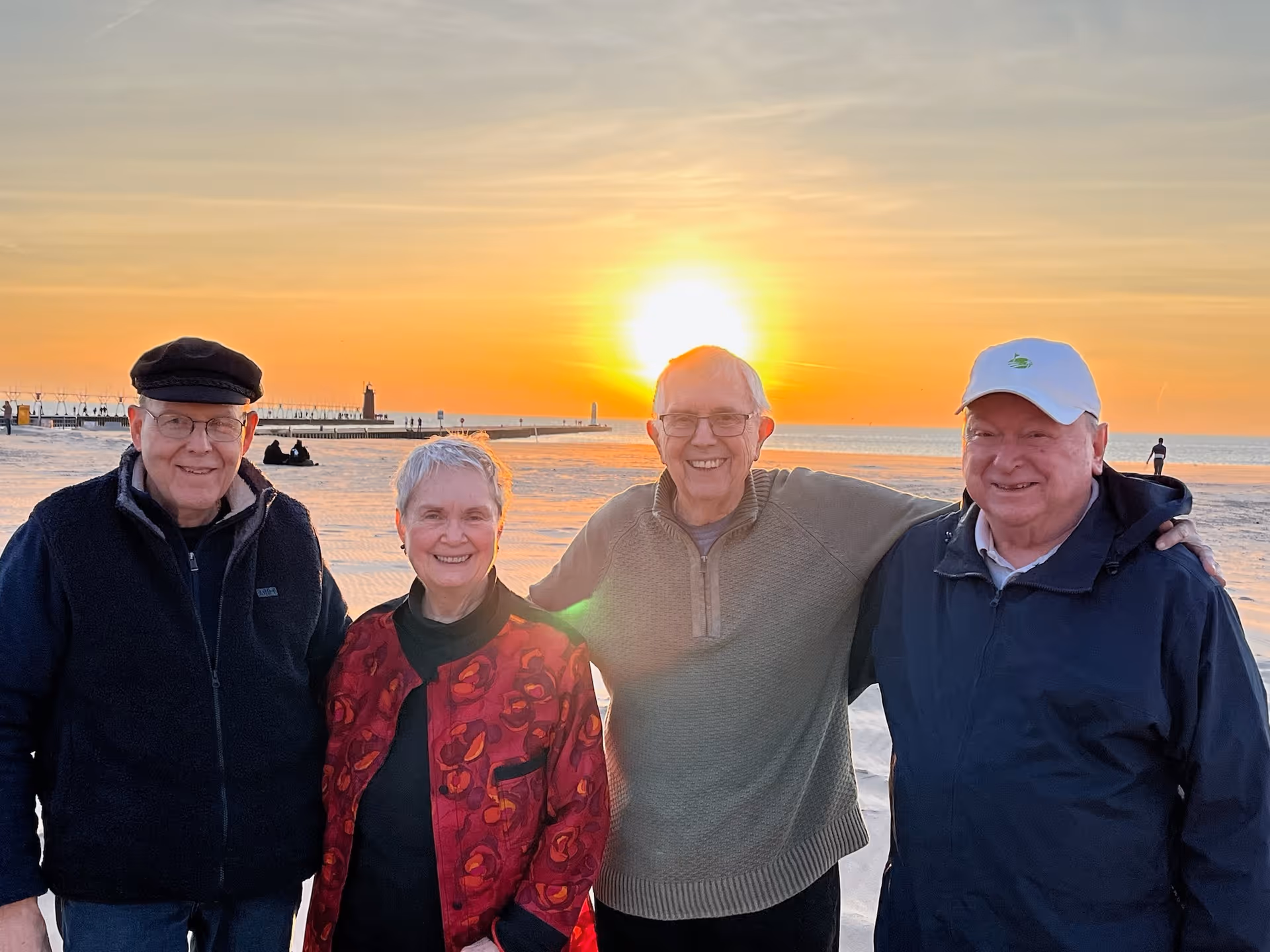 Four elderly adults standing close together on a beach at sunset, smiling at the camera. The sun is low on the horizon over the water, casting a warm glow. In the background, there is a pier with a lighthouse and a few people walking along the shore.