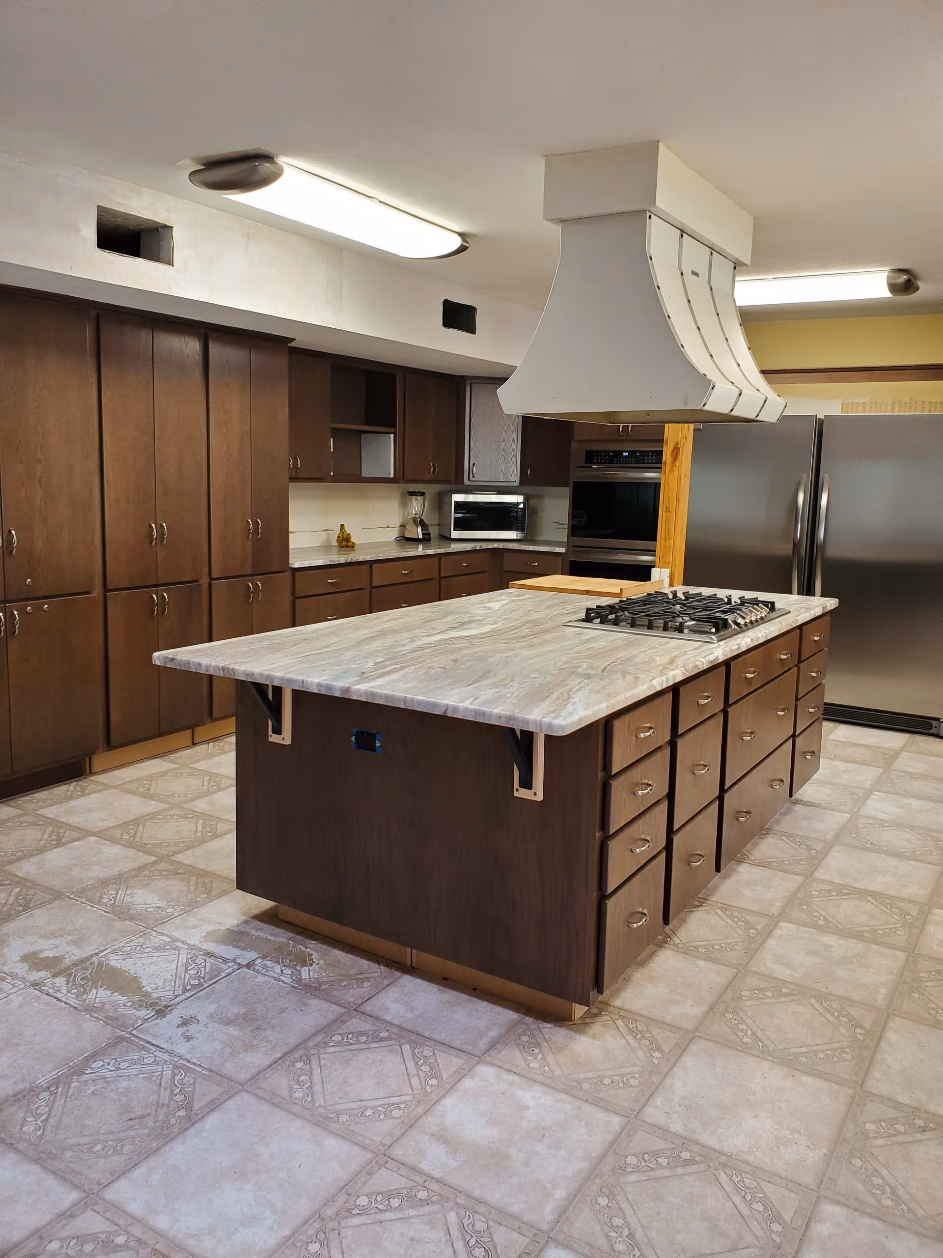 A spacious kitchen with a large central island featuring a built-in gas stove and multiple drawers. The kitchen has dark wooden cabinets along the walls, a double-door stainless steel refrigerator, a built-in oven, a microwave, and a blender on the countertop. The floor is tiled with a patterned design, and there are two ceiling lights providing illumination.