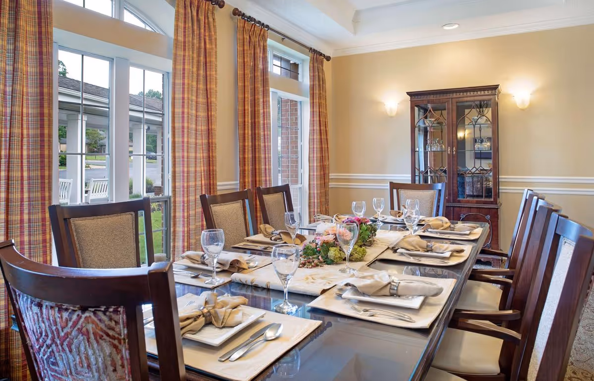 A formal dining room with a long glass-top table set for eight people. Each place setting includes a plate, napkin with a napkin ring, fork, knife, spoon, and a wine glass. The room has large windows with plaid curtains, beige walls with white trim, and a wooden china cabinet in the corner displaying glassware. Soft wall sconces provide lighting.