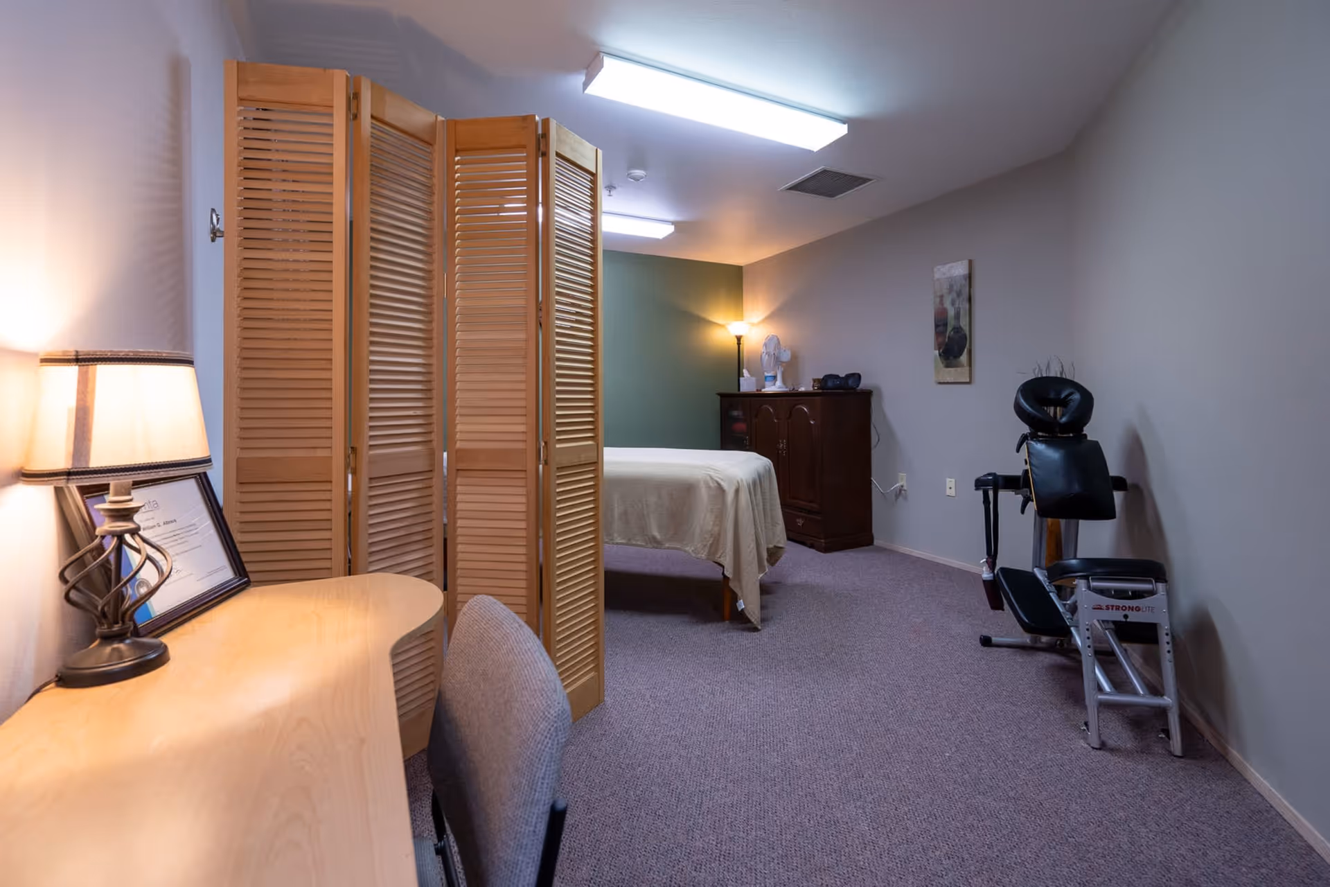 A quiet room with a massage table covered in a beige sheet, a wooden folding screen, a wooden desk with a lamp and framed certificate, a gray chair, a dark wooden cabinet with a fan and other items on top, a floor lamp, a piece of wall art, and a black exercise machine against a gray wall.