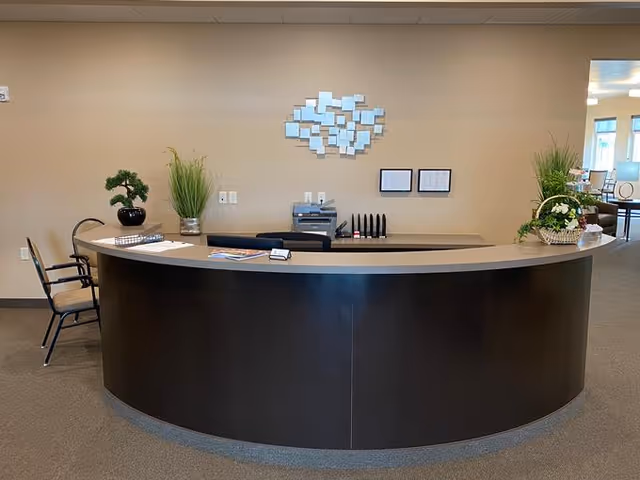 A curved reception desk in an office or facility lobby with two chairs on the left side, a small potted plant, a vase with tall grass, and office supplies on the desk. Behind the desk, there is a printer, a wall decoration made of small square mirrors, and two framed documents on a beige wall. To the right, there is an open doorway leading to another room with chairs and a table.