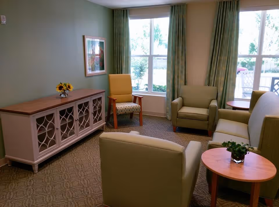 A cozy living room with green walls and carpet, featuring a wooden sideboard with a vase of sunflowers, a framed picture on the wall, two green armchairs, a green sofa, a yellow armchair with wooden arms, two round wooden side tables, and large windows with green curtains letting in natural light.