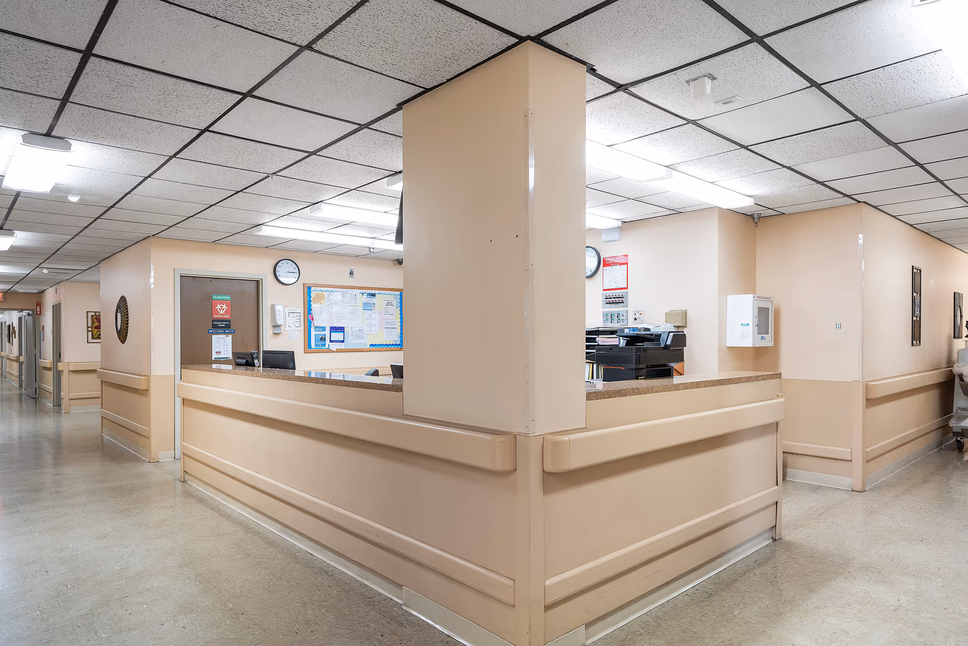 Interior view of a nursing center hallway with a central nurse's station featuring beige walls, a bulletin board, clocks, and office equipment. The hallway extends on both sides with handrails along the walls and fluorescent ceiling lights.