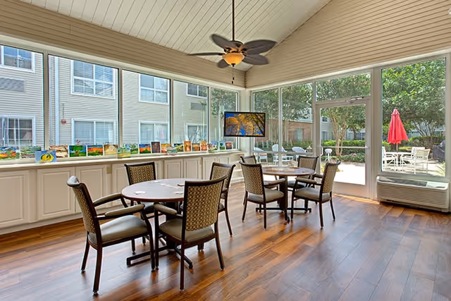 A bright and spacious common area with large windows showing an outdoor patio. The room has wooden floors, a ceiling fan with a light, and several round tables with cushioned chairs arranged around them. Colorful artwork is displayed on a long white cabinet beneath the windows.