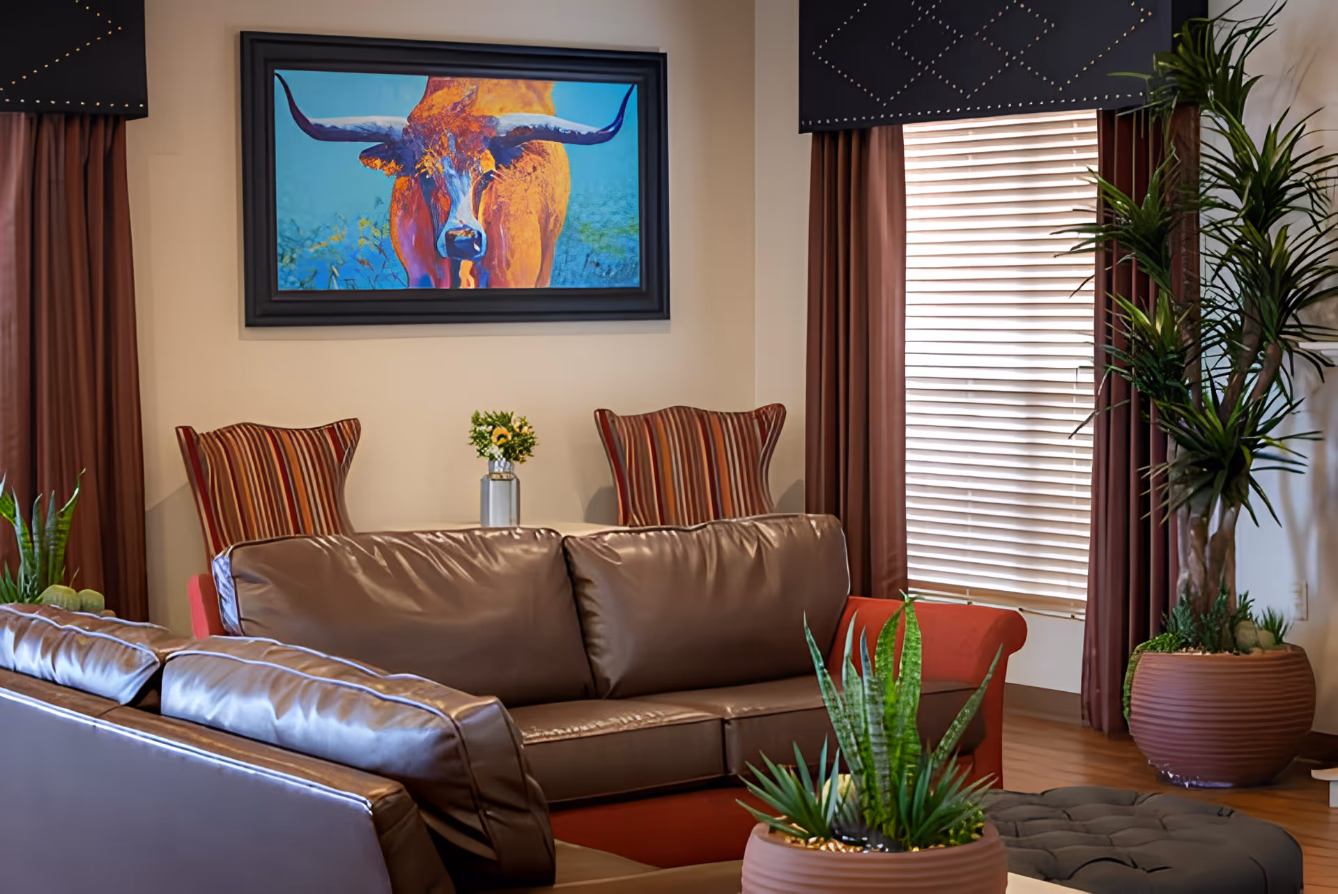A cozy living room area with brown leather sofas, two striped armchairs, a large potted plant, and a window with blinds and brown curtains. A colorful painting of a longhorn bull is hung on the wall above the armchairs.