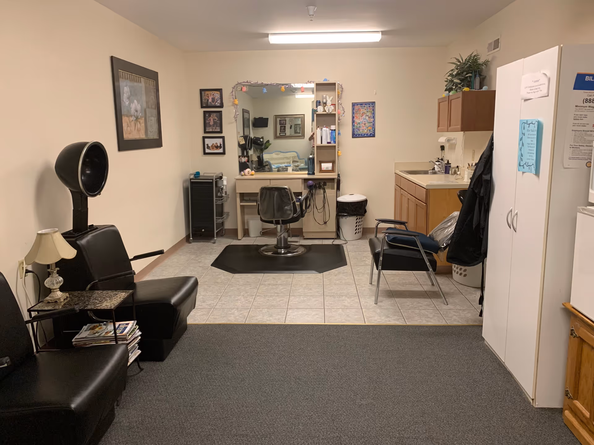 Interior view of a small salon area in South Pointe Senior Apartments featuring a black salon chair with a hair dryer hood, a vanity mirror with shelves and a salon chair in front, a small sink and cabinet area, two black chairs for waiting, a small table with a lamp and magazines, and various decorations on the walls.
