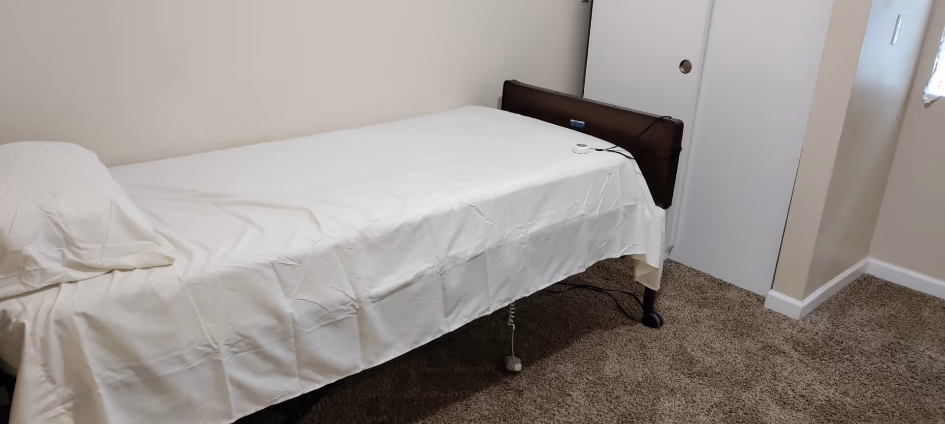 A small bedroom with a single bed covered in white sheets, beige walls, and brown carpet.
