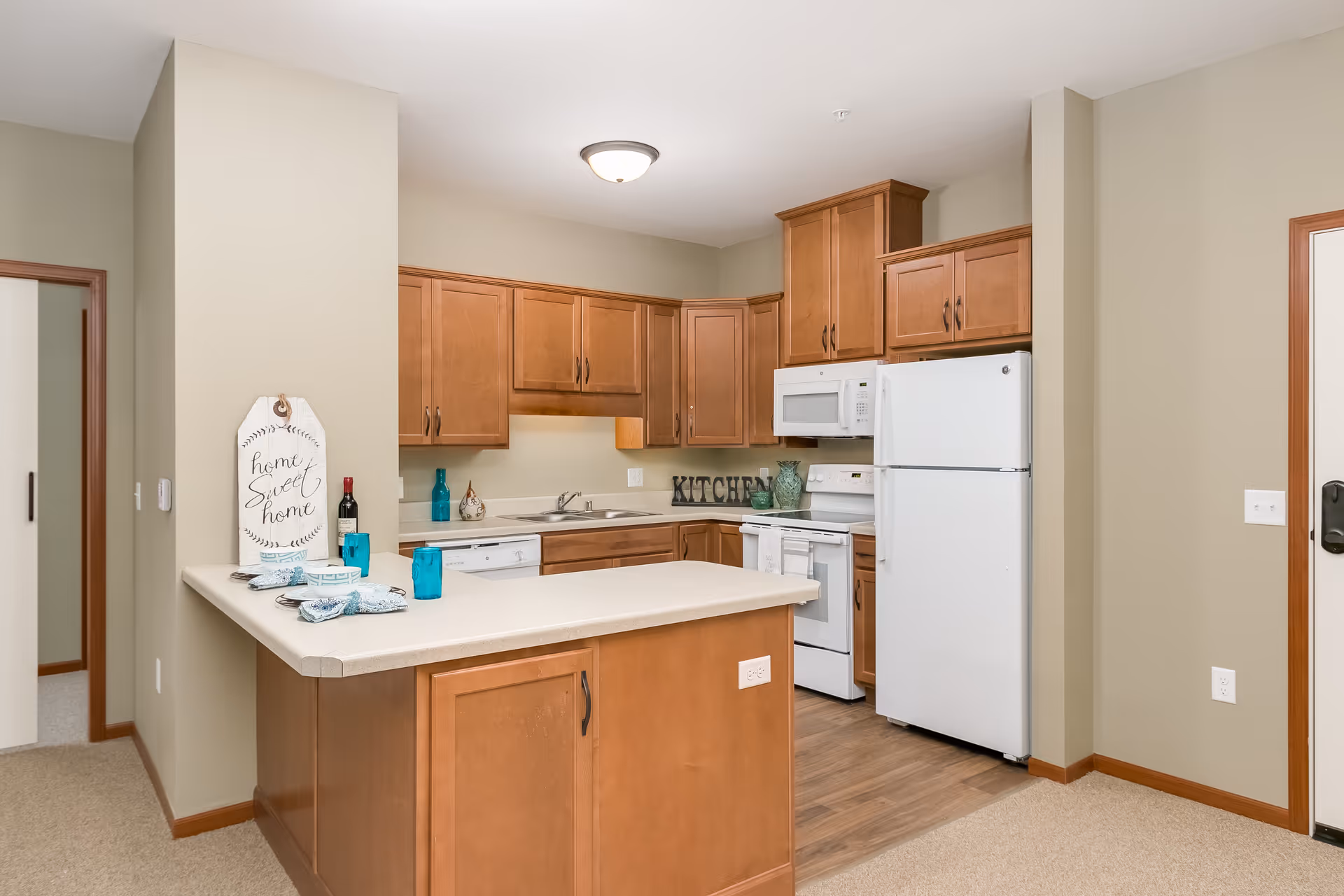 A clean and modern kitchen with wooden cabinets, a white refrigerator, microwave, stove, and dishwasher. There is a countertop island with place settings including plates, bowls, and blue glasses. The walls are painted beige and the floor is a combination of carpet and wood.