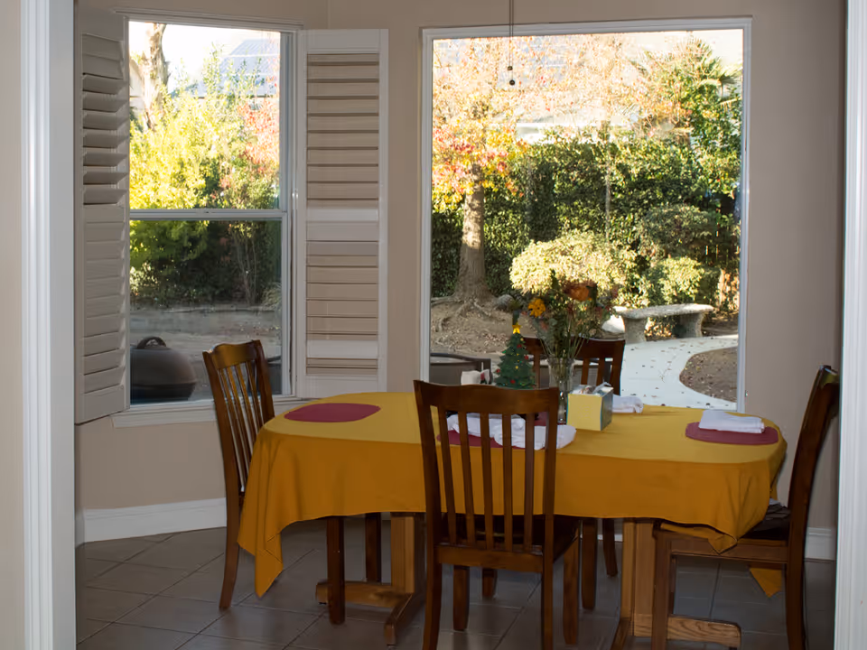 A dining area with a wooden table covered by a yellow tablecloth and four wooden chairs. The table is set with placemats, folded napkins, a tissue box, and a vase of flowers. Large windows behind the table show a garden with trees, bushes, a stone bench, and a curved pathway.