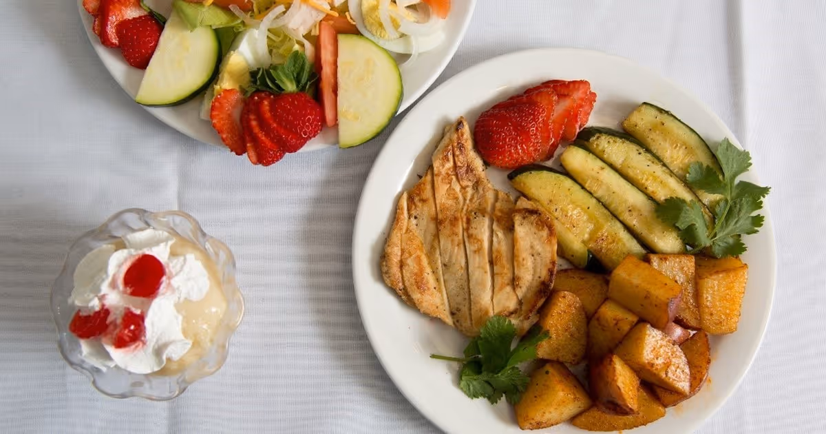 Plate of grilled chicken with roasted potatoes, zucchini and strawberries alongside a salad and a dessert cup on a white tablecloth.