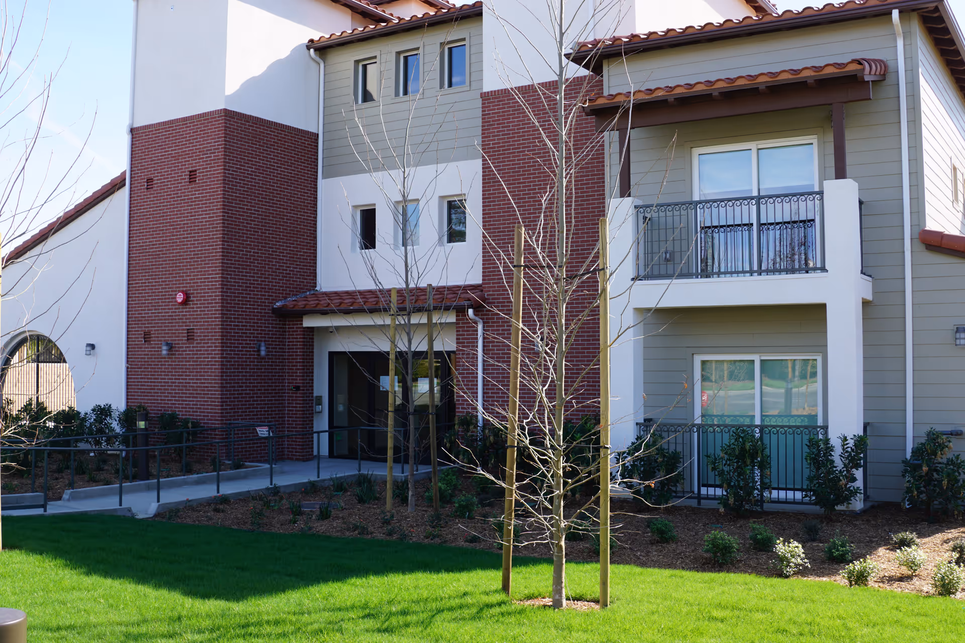 Exterior view of a modern residential building with a combination of brick and siding walls, featuring balconies with metal railings, a green lawn, and young trees supported by wooden stakes in the foreground.