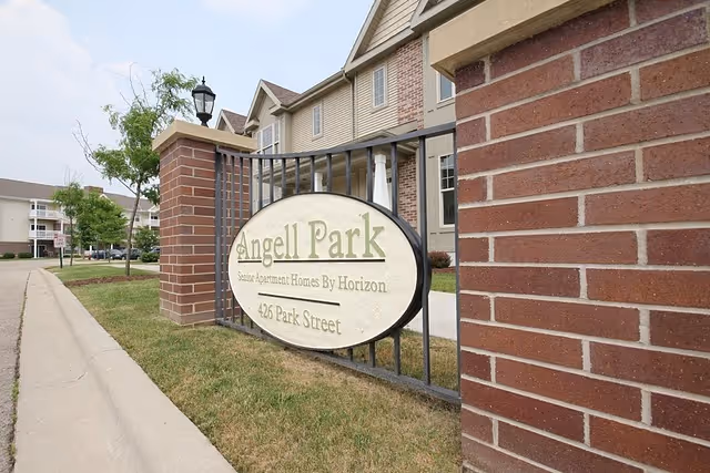 Entrance sign for Angell Park senior apartments mounted on a fence in front of a brick apartment building.