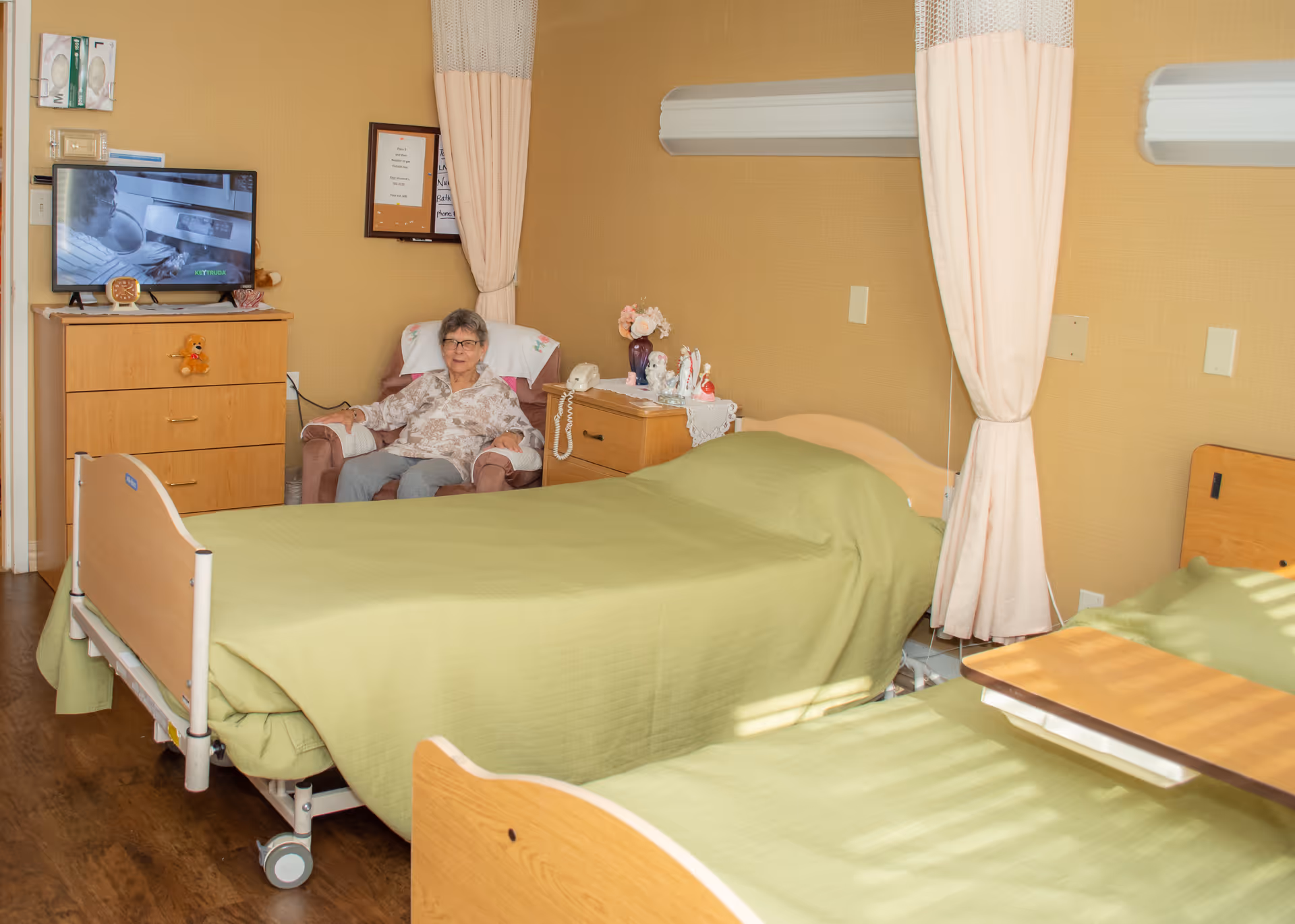 A nursing home bedroom with two hospital beds covered in green blankets and an elderly woman seated in a chair beside a dresser and TV.