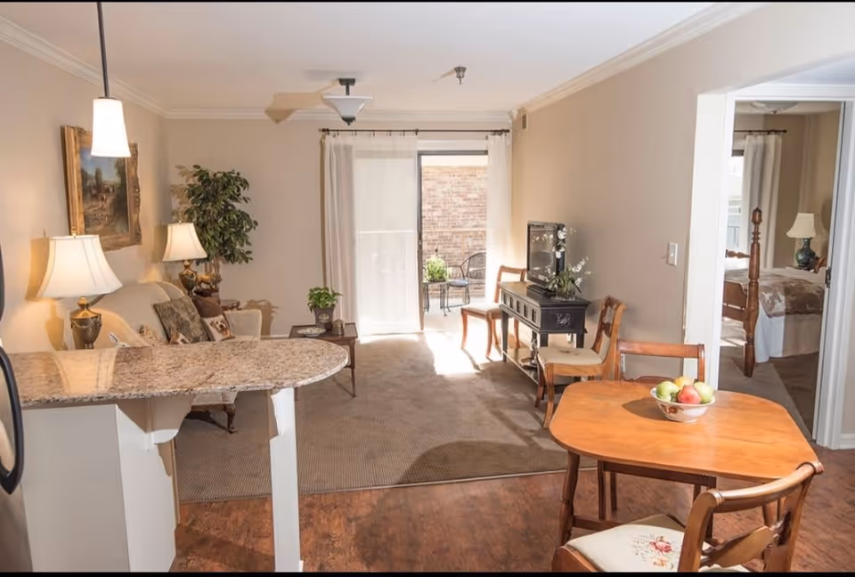 A cozy living area in an assisted living apartment featuring a small round wooden dining table with four chairs, a granite countertop bar, a beige sofa with decorative pillows, two table lamps, a potted plant, a TV on a black stand, and a sliding glass door leading to a small outdoor patio with chairs. A bedroom with a wooden bed frame and a lamp is visible through an open doorway.