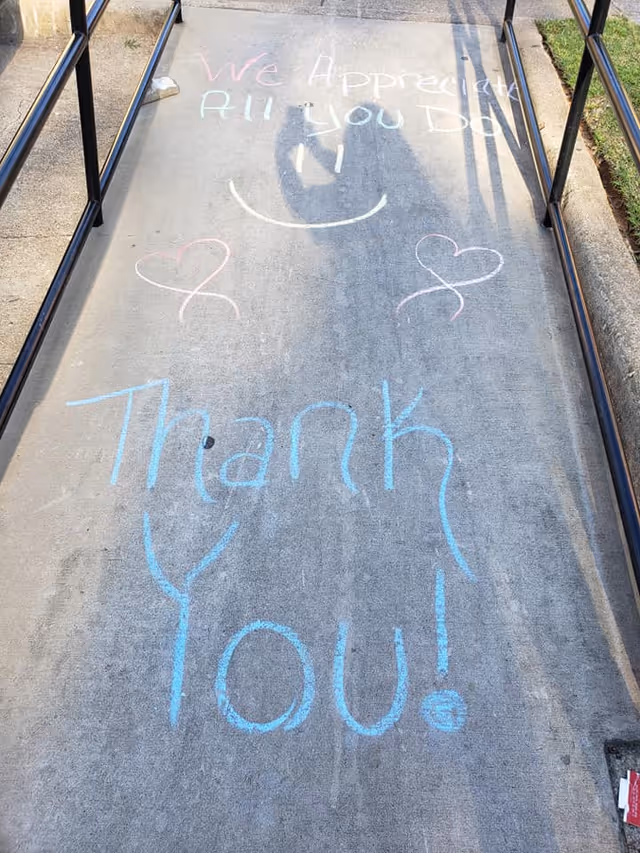 Sidewalk ramp with metal railings on both sides featuring colorful chalk writing that says 'We Appreciate All You Do :)' with two hearts and 'Thank You!'