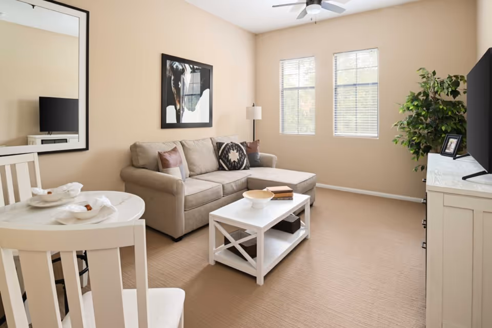 Bright furnished living room with a beige sectional sofa, white coffee table, small dining table and TV.