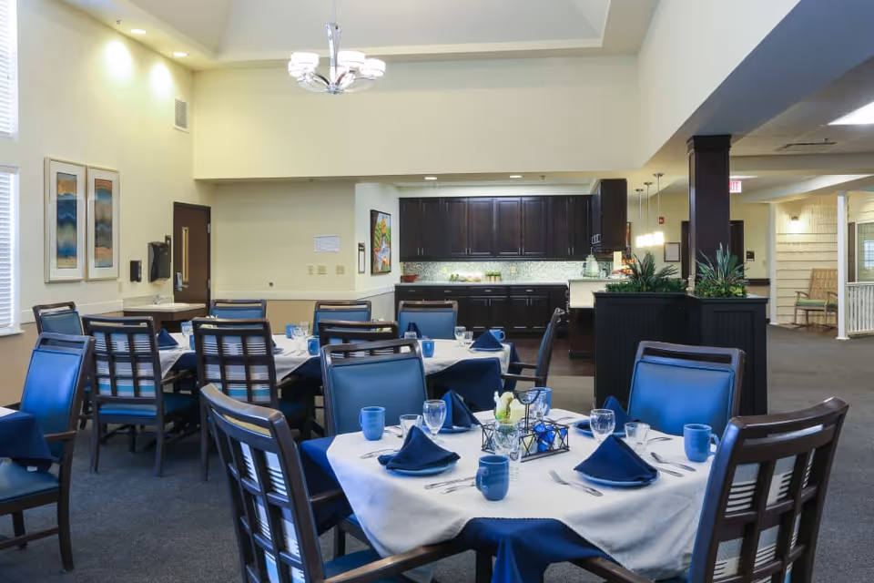 A dining room in Orchard Grove Senior Living with several tables set for a meal. Each table has a white tablecloth with a blue overlay, blue napkins, blue mugs, and glassware. The room features dark wooden chairs with blue cushions, light yellow walls, framed artwork, and a kitchen area with dark cabinets in the background. There are plants on a divider and ceiling lights providing illumination.