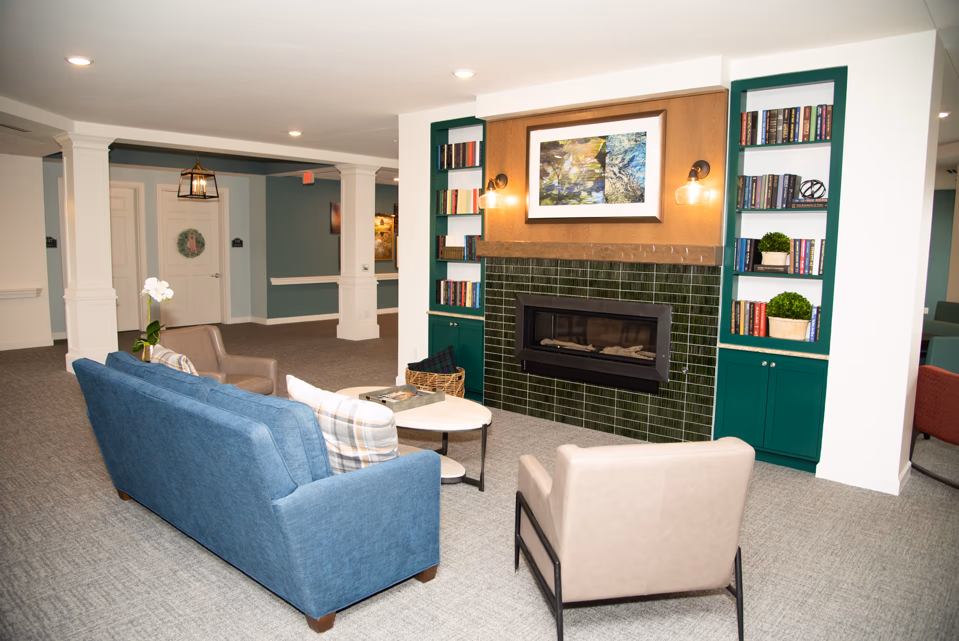 A cozy seating area in a senior living facility with a blue sofa, two beige armchairs, and a round coffee table. The background features a fireplace with green tiles, built-in bookshelves on either side filled with books and decorative plants, and a framed artwork above the fireplace. The room has light-colored walls and carpeted floors, with white columns and doors visible in the hallway.