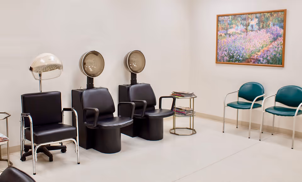 Interior of a salon area with three black salon chairs, two with attached hair dryers and one with a hooded hair dryer. There are two green chairs against the wall, a small table with magazines, and a framed painting of a colorful garden scene on the wall.