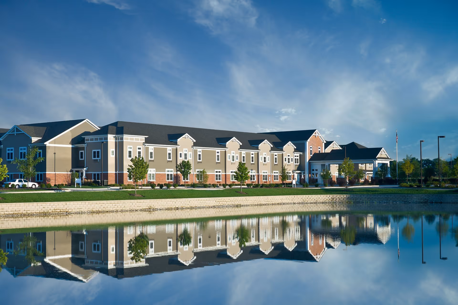 A large, modern senior living facility building with beige and brick exterior walls, multiple windows, and a dark roof, reflected in a calm pond in front of it under a partly cloudy blue sky.
