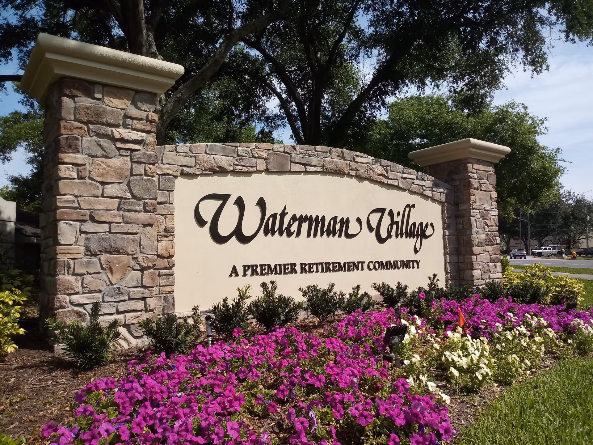 Stone and stucco sign for Waterman Village, a premier retirement community, surrounded by vibrant purple and white flowers and greenery with trees and a road in the background.