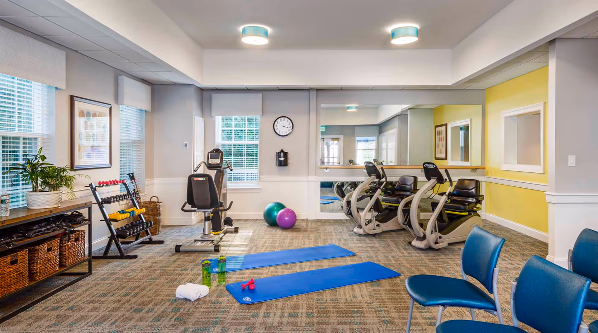 A well-lit fitness room with exercise equipment including two recumbent bikes, a weight rack with dumbbells, two exercise mats on the carpeted floor, two exercise balls, and several blue chairs. The room has large windows with blinds, a wall clock, a large mirror, and light yellow and gray walls.