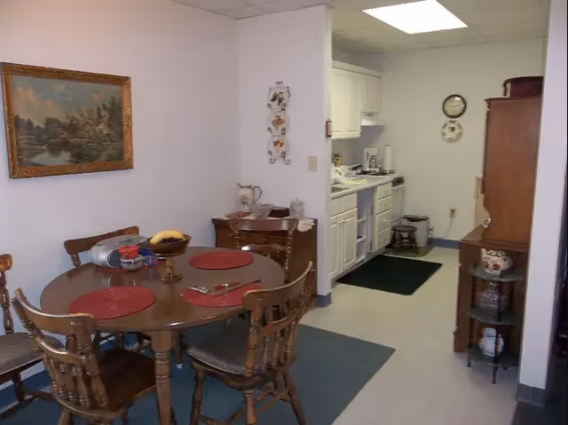 A small dining area with a round wooden table set with four chairs and red placemats. The table has a fruit bowl and some other items on it. Adjacent to the dining area is a compact kitchen with white cabinets, a stove, and a small stool. The walls are white, and there is a framed landscape painting on the wall above the dining table. A wooden cabinet and a small round shelf with decorative items are also visible.
