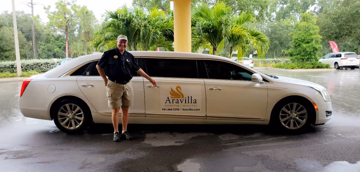 A man wearing a black shirt, khaki shorts, and a cap stands next to a white limousine with a sign on the door that reads 'Aravilla Assisted Living | Memory Care 941.444.9398 | Aravilla.com'. The scene is outdoors with palm trees and other greenery in the background on a rainy day.