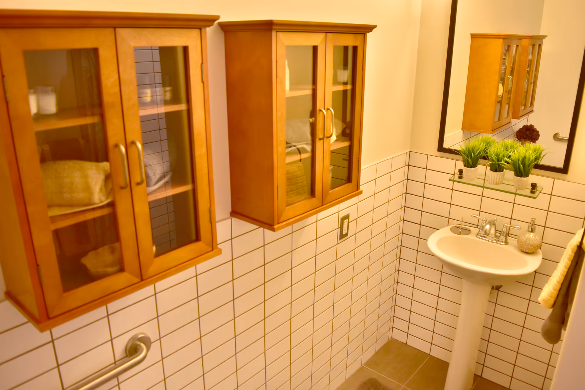 A small bathroom with white tiled walls and a pedestal sink. Above the sink is a mirror and a glass shelf holding three small potted plants. Two wooden cabinets with glass doors are mounted on the wall to the left. A handrail is visible on the left side wall, and a towel hangs on the right side.