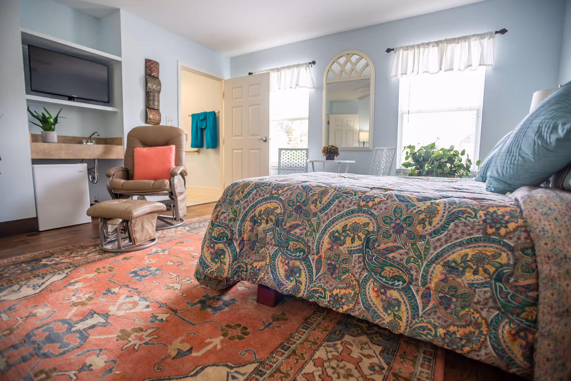 Sunlit bedroom with a patterned bedspread, reclining armchair and ottoman, wall-mounted TV, and windows.