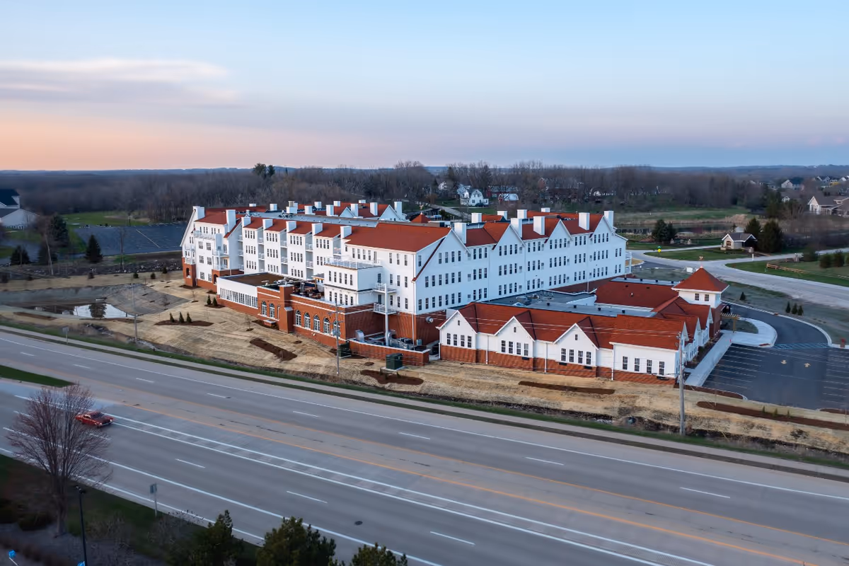 Aerial view of The Westerly Pewaukee, a large multi-story senior living facility with white walls and red roofs, situated next to a wide road with a few cars and surrounded by open land and trees.