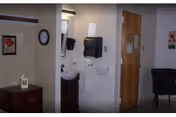 Interior view of a room in a senior living facility showing a small sink area with a mirror, soap dispenser, and paper towel holder mounted on the wall. To the left, there is a small wooden cabinet with a decorative item on top and a framed picture on the wall. A clock is also visible on the wall above the cabinet. To the right, there is a wooden door and a dark upholstered chair against the wall.