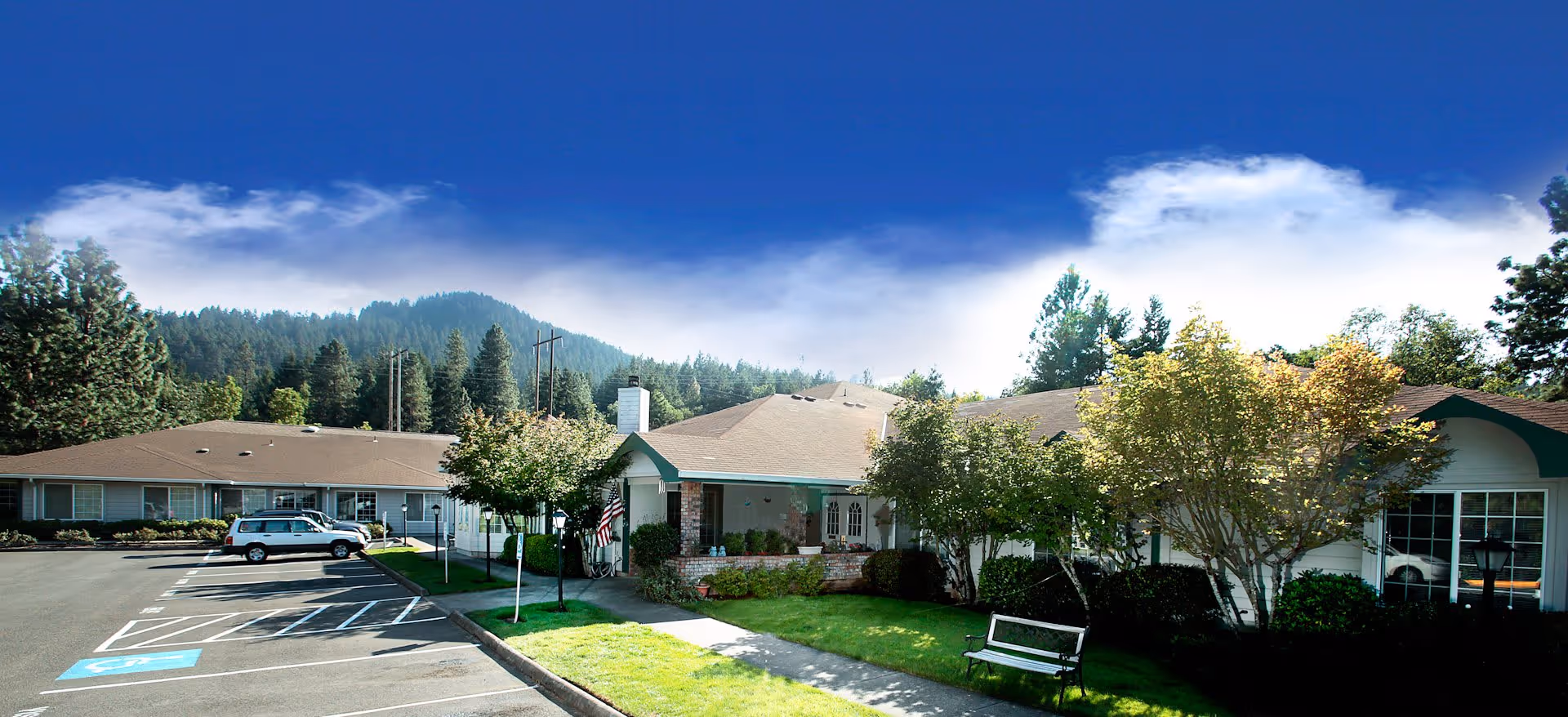Exterior view of Fox Hollow Senior Living facility showing a single-story building with a parking lot, green lawn, trees, and a bench under a bright blue sky with some clouds and forested hills in the background.