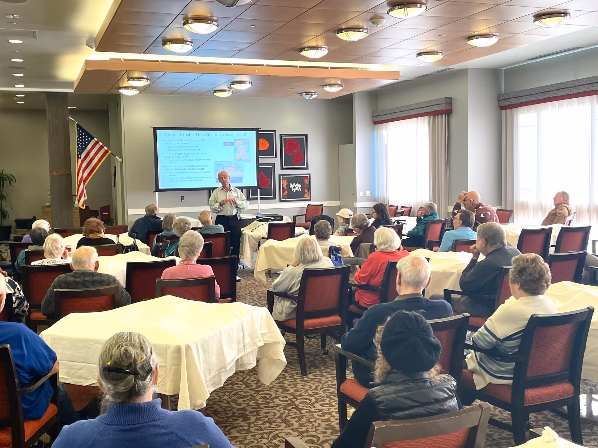 A group of elderly people seated in a community room attentively listening to a man giving a presentation. The presenter stands near a screen displaying slides about nuclear weapons history. The room has tables covered with white tablecloths, red cushioned chairs, large windows with curtains, and an American flag in the corner.