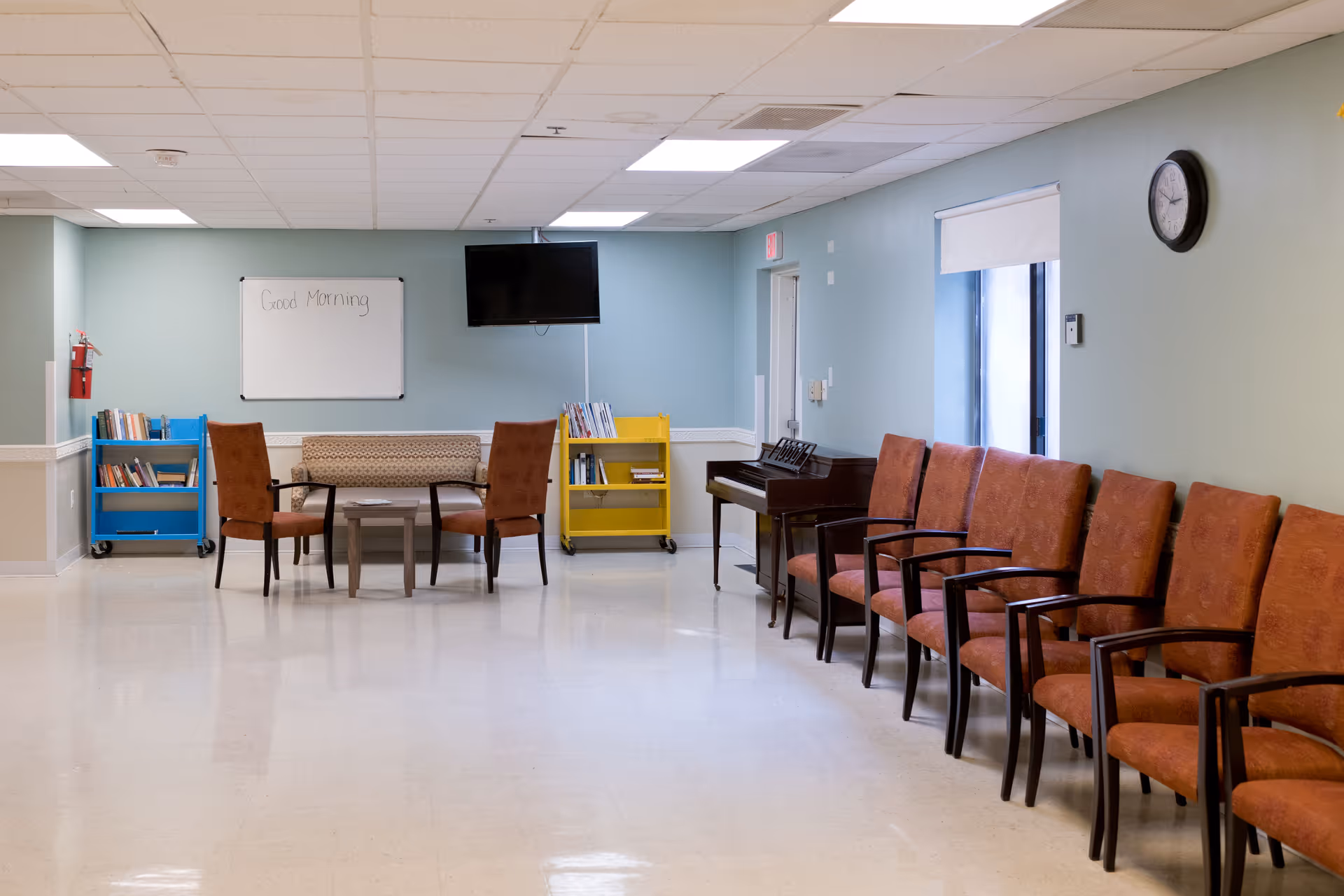A spacious room with a row of brown chairs lined up against a light blue wall. There is a small seating area with two brown chairs, a small table, and a patterned bench. Two book carts, one blue and one yellow, are filled with books. A whiteboard on the wall says 'Good Morning'. A flat-screen TV is mounted above the whiteboard. A piano is positioned near a window with a clock above it on the wall.