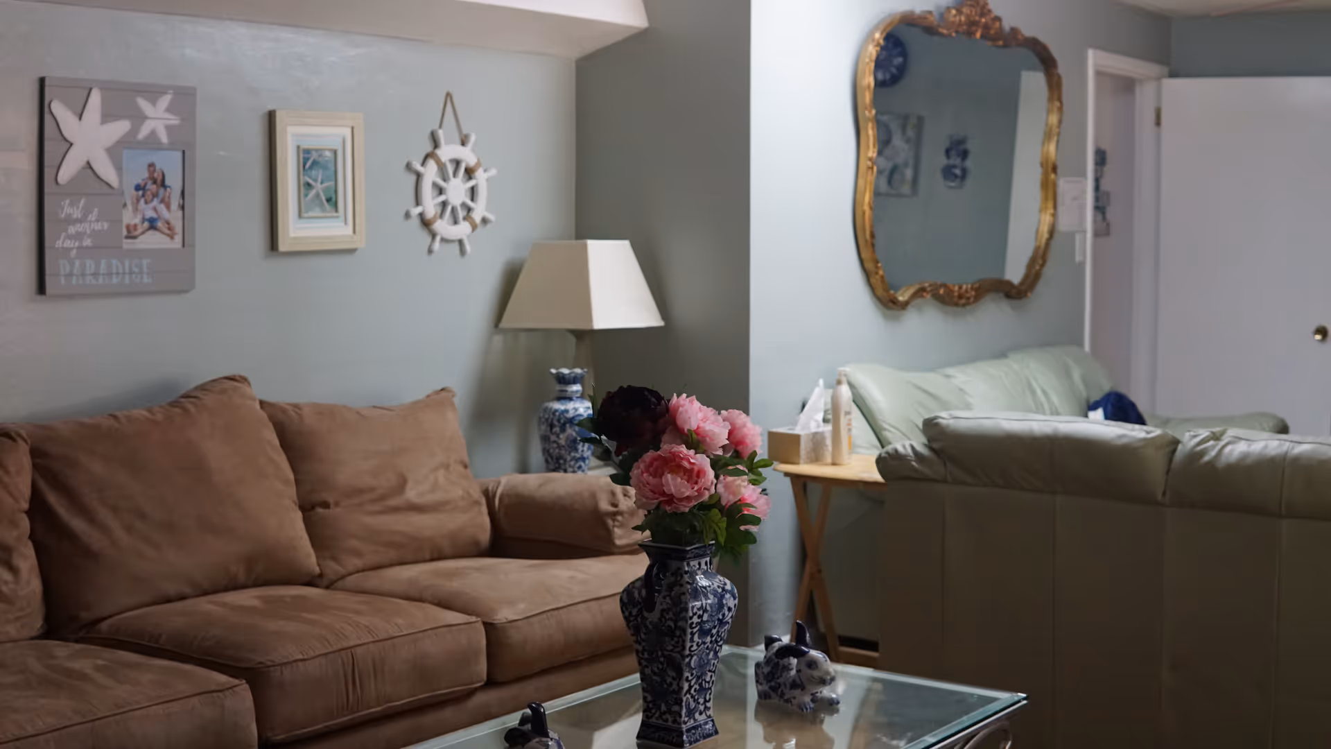 A cozy living room with a brown sofa, a glass coffee table topped with a blue-and-white vase of pink flowers, wall decor and a gold-framed mirror.