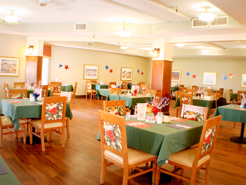 A bright dining room with multiple tables covered in green tablecloths and wooden chairs with floral upholstery. The tables are set with utensils, napkins, and decorative centerpieces. The room has wooden flooring, light-colored walls adorned with framed pictures and red and blue star decorations, and ceiling lights providing warm illumination.
