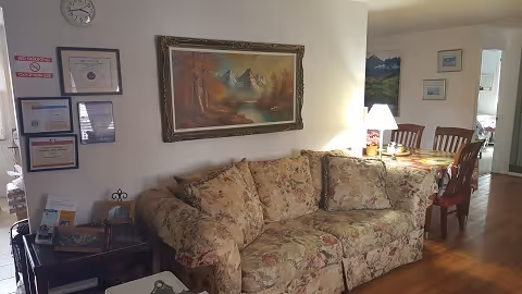 A cozy living room area with a floral patterned sofa against a wall. Above the sofa hangs a framed painting of a mountain landscape. To the right, there is a dining area with a wooden table and chairs, a table lamp, and some framed pictures on the wall. Certificates and signs are displayed on the wall to the left of the sofa. The floor is wooden, and the room is softly lit.