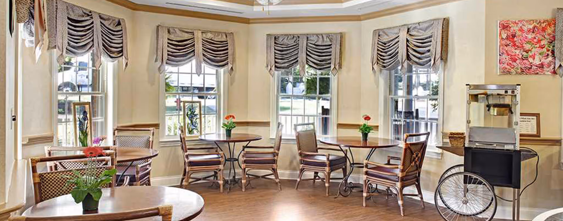 Bright dining/lounge area with round tables and chairs, potted flowers on the tables, large windows with valances, and a popcorn cart.