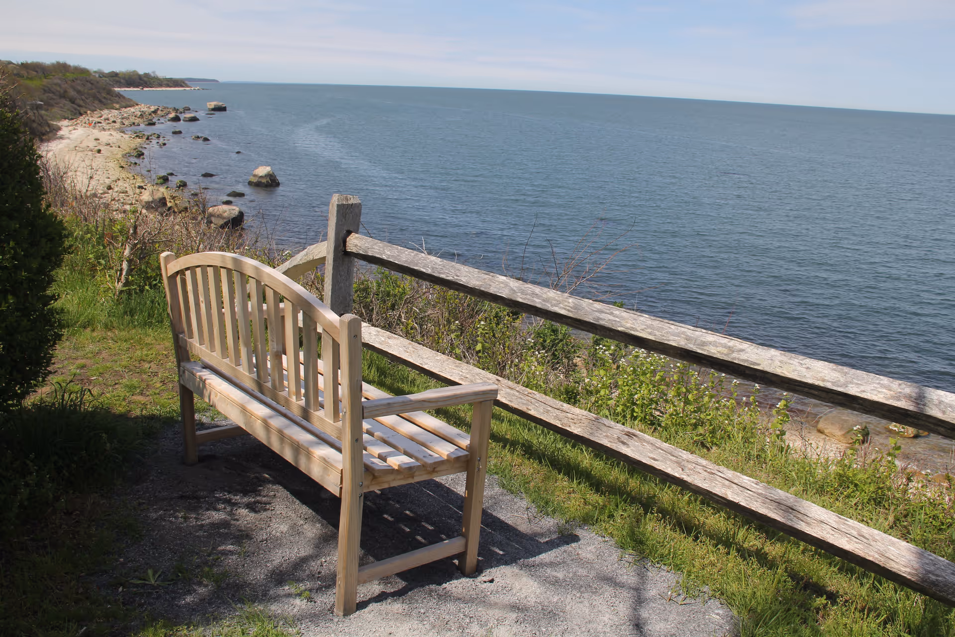 A wooden bench facing a large body of water with a rocky shoreline and greenery. A wooden fence runs parallel to the water, separating the bench area from the edge of the shore under a clear sky.